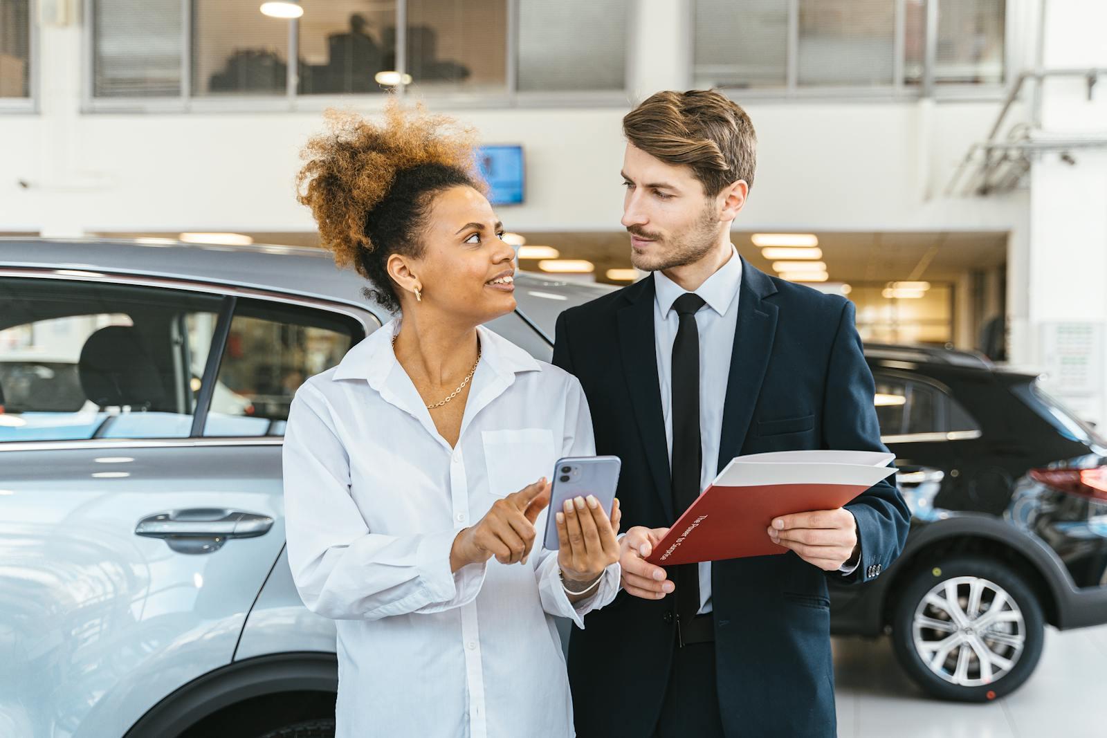 Une femme afro-américaine et un homme caucasien discutent de l'achat d'une voiture chez un concessionnaire à l'aide d'un smartphone.